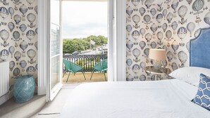 A bedroom with a balcony, patterned wallpaper, a bed with a blue headboard, and a small table with a lamp. - The Zetland Apartment (Saltburn-by-the-Sea)