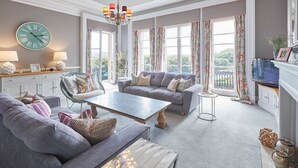 A spacious living room with a grey sofa, a coffee table, and a TV. There are large windows with curtains, a chandelier, and a clock on the wall. - The Zetland Apartment (Saltburn-by-the-Sea)
