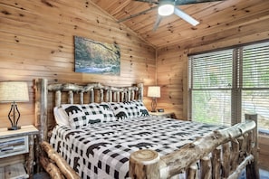 A wooden cabin bedroom with a bed, two nightstands, a ceiling fan, and a window with blinds.