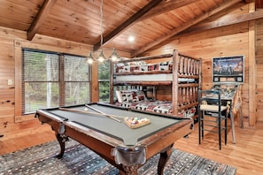 A pool table with a rack of billiard balls, a sofa with patterned cushions, and a wooden bookshelf with various items.