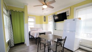 A compact room with a bed, a dining table with chairs, a ceiling fan, a refrigerator, and a television on the wall. - Studio w/ Kitchenette 2 Blocks to Beach & Main Ave | Near Asbury Boardwalk (Ocean Grove)