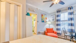 Un dormitori amb llit, ventilador de sostre, finestra amb cortines, porta amb el símbol de número i taula petita amb cadira.