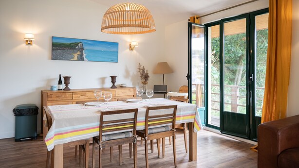 Un coin repas avec une table en bois, des chaises et une nappe à rayures. Il y a un tableau au mur, une lampe et une fenêtre avec des rideaux.