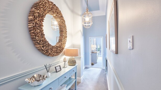 A hallway with a light blue wall, a decorative wooden wreath on the wall, a light blue console table with a white bowl, a lamp, and a picture frame. - The Zetland Apartment (Saltburn-by-the-Sea)