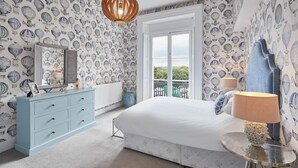 A bedroom with a large bed, a dresser, a round table with a lamp, and a view of the outdoors through a window. - The Zetland Apartment (Saltburn-by-the-Sea)
