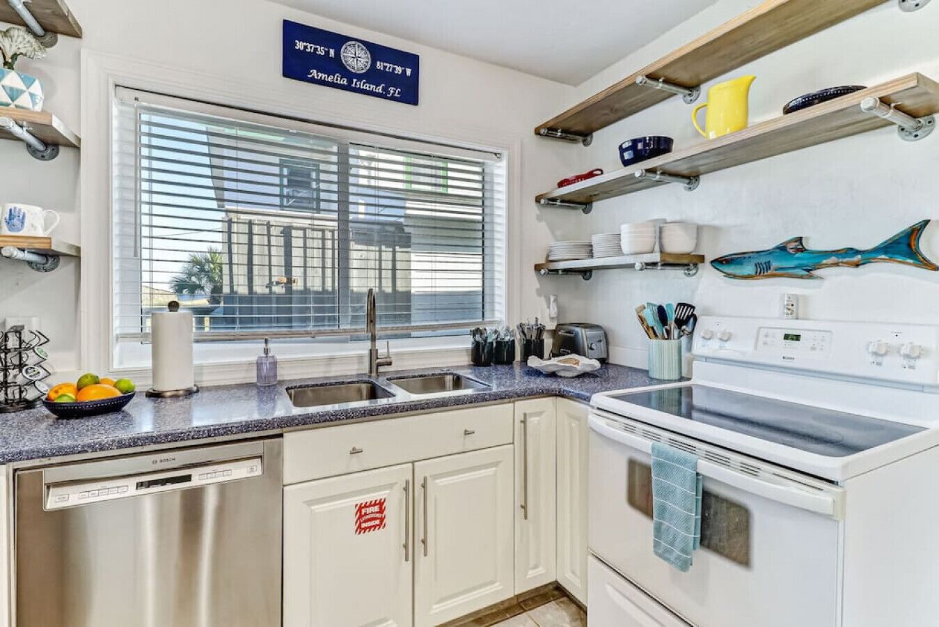 A kitchen with a built-in dishwasher, a stove, and a sink. There is a window with blinds, a shelf with kitchen utensils, and a sign indicating the location.