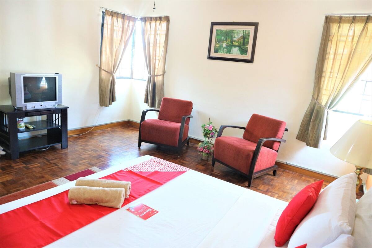 A hotel room with a bed, two red chairs, a TV on a stand, and a painting on the wall.
