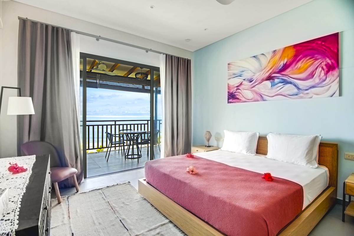 Ein Hotelzimmer mit Bett, Schreibtisch, Stuhl, einem Balkon mit Aussicht und einem Bild an der Wand.