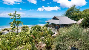 A coastal property with a large open deck overlooking the ocean, surrounded by lush greenery and a clear blue sky.