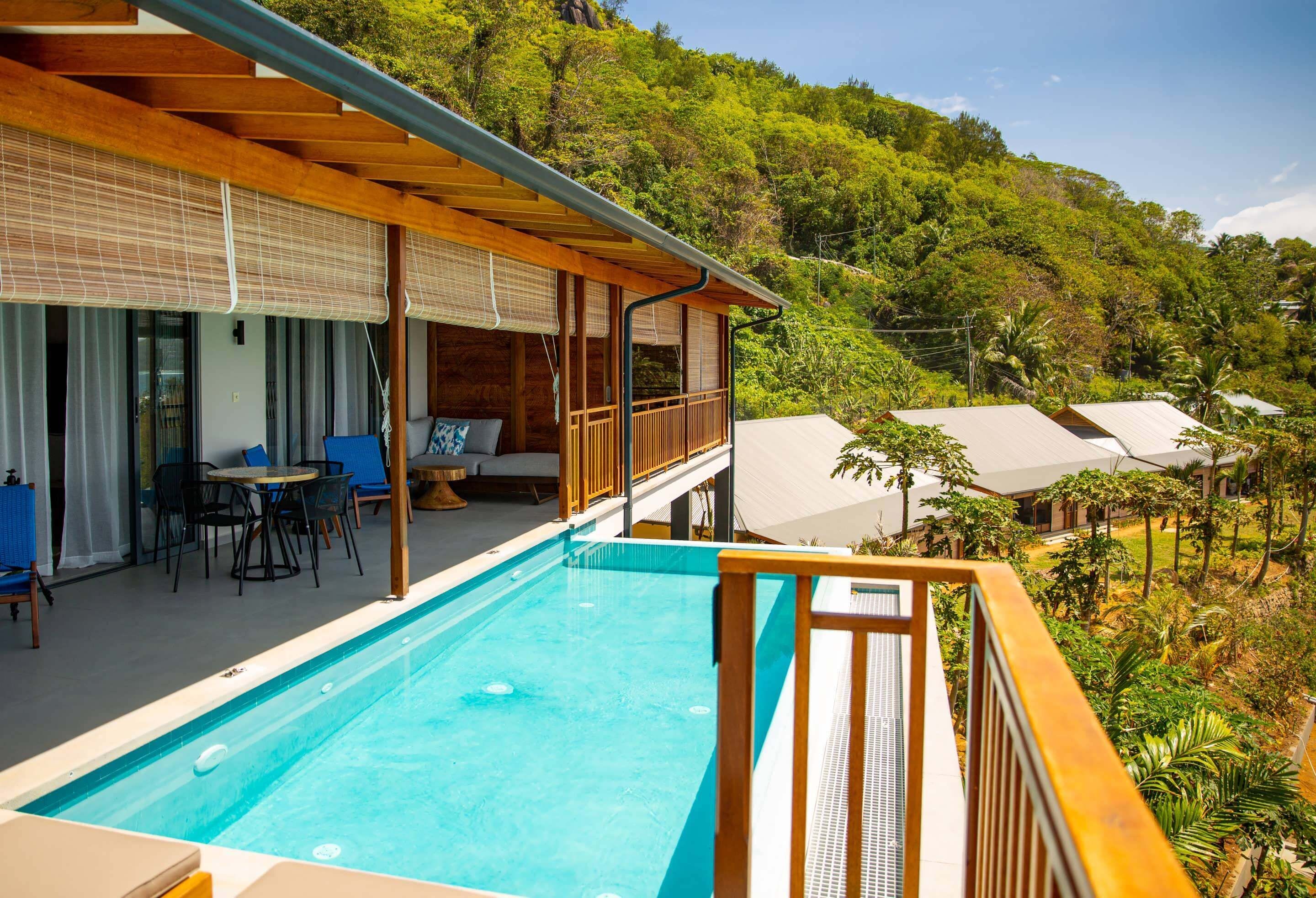 A poolside area with a wooden deck, a covered seating area, and a view of a lush green hillside.