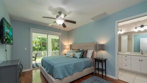 Un dormitorio con cama, ventilador de techo, televisor, baño con espejo e iluminación, y vistas al exterior con vegetación.