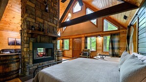 Ein Schlafzimmer in einer Blockhütte mit Kamin aus Stein, einem großen Bett und Blick ins Freie durch die Fenster.