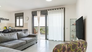 A modern living room with a grey sofa, a patterned armchair, a kitchen area with a sink and counter, and a sliding glass door leading to a balcony.