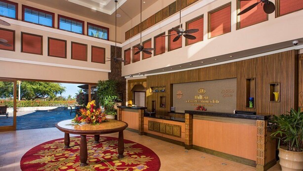 Un espace d’accueil d’hôtel comprenant un comptoir de réception, une table ronde surmontée d’un tapis à motifs floraux, des ventilateurs de plafond et une vue sur l’espace extérieur.