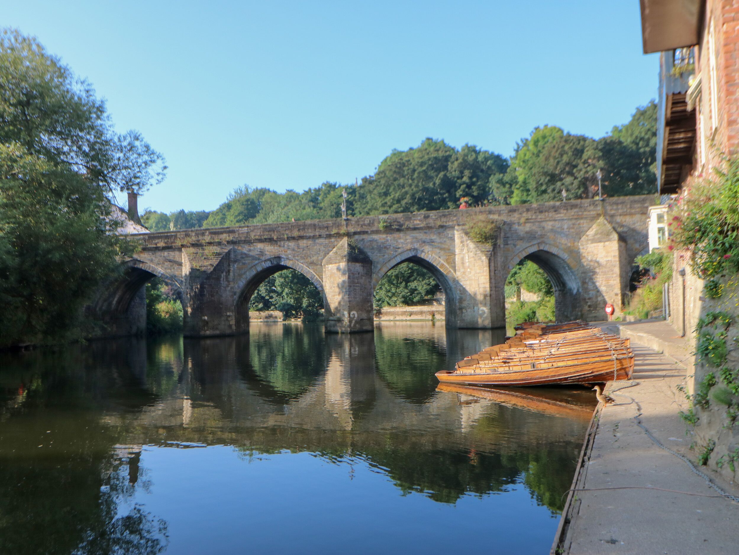 A stone bridge over a calm river with a wooden boat docked on the right.