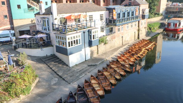 Un bâtiment situé au bord de la rivière, avec un quai où sont amarrés des bateaux en bois.