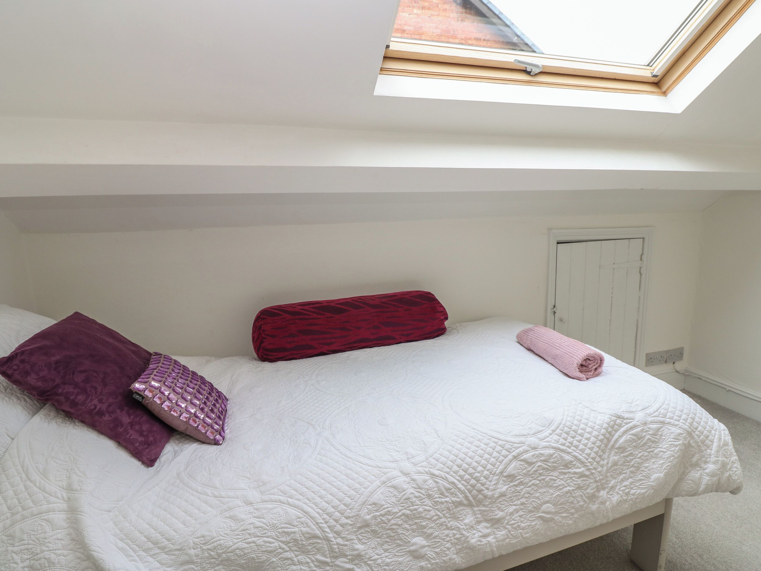 A bedroom with a skylight, a bed with a white quilt, a red pillow, and a purple pillow.