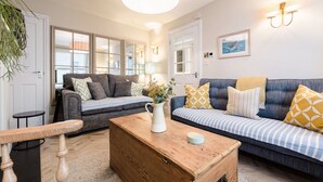 A living room with a wooden coffee table, two sofas with patterned cushions, a hanging light fixture, and a view of the outdoors through large windows.