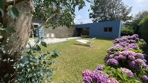 Un jardin avec une pelouse, un arbre, un bâtiment moderne de couleur bleue et des fleurs d’hortensia violettes.