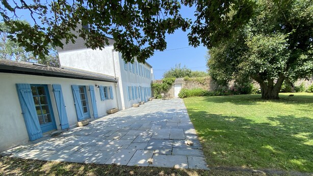 A paved pathway leading to a white building with blue shutters, surrounded by greenery and a large tree.