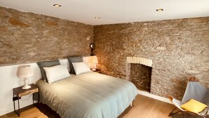 Une chambre avec un mur en pierre, un lit recouvert d’une couverture grise, une table de chevet avec une lampe, une chaise et une cheminée.