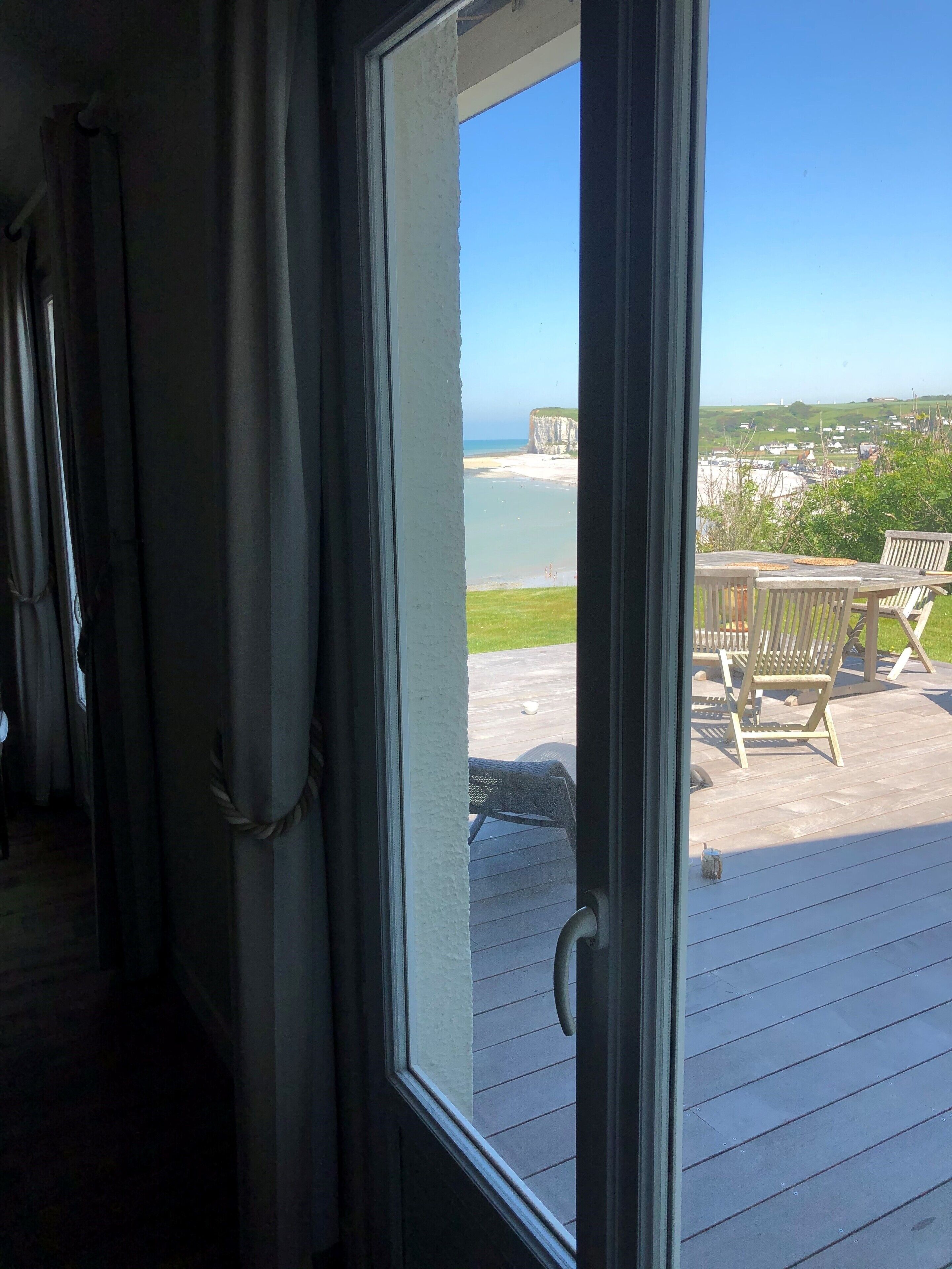 Blick aus einem Zimmer mit großem Fenster auf eine Terrasse mit Gartenmöbeln und einen Strand.