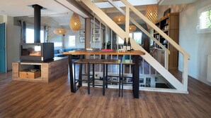 Un vaste espace de vie doté d’un plancher en bois, d’une table centrale entourée de chaises, d’un escalier et d’une bibliothèque.