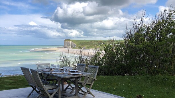 Une terrasse en bois avec une table et des chaises, donnant sur une plage et des falaises.