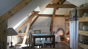 Une chambre mansardée chaleureuse avec un plafond en pente, des poutres en bois, un lit, une table avec des chaises et une étagère remplie de vaisselle.