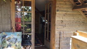 Une cabane en bois avec une grande porte ouverte donnant sur un couloir. On y trouve une petite table recouverte d’une nappe colorée et une chaise.