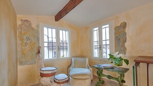 Una habitación con un ventanal grande, una silla, una mesita con un florero y un taburete decorativo.