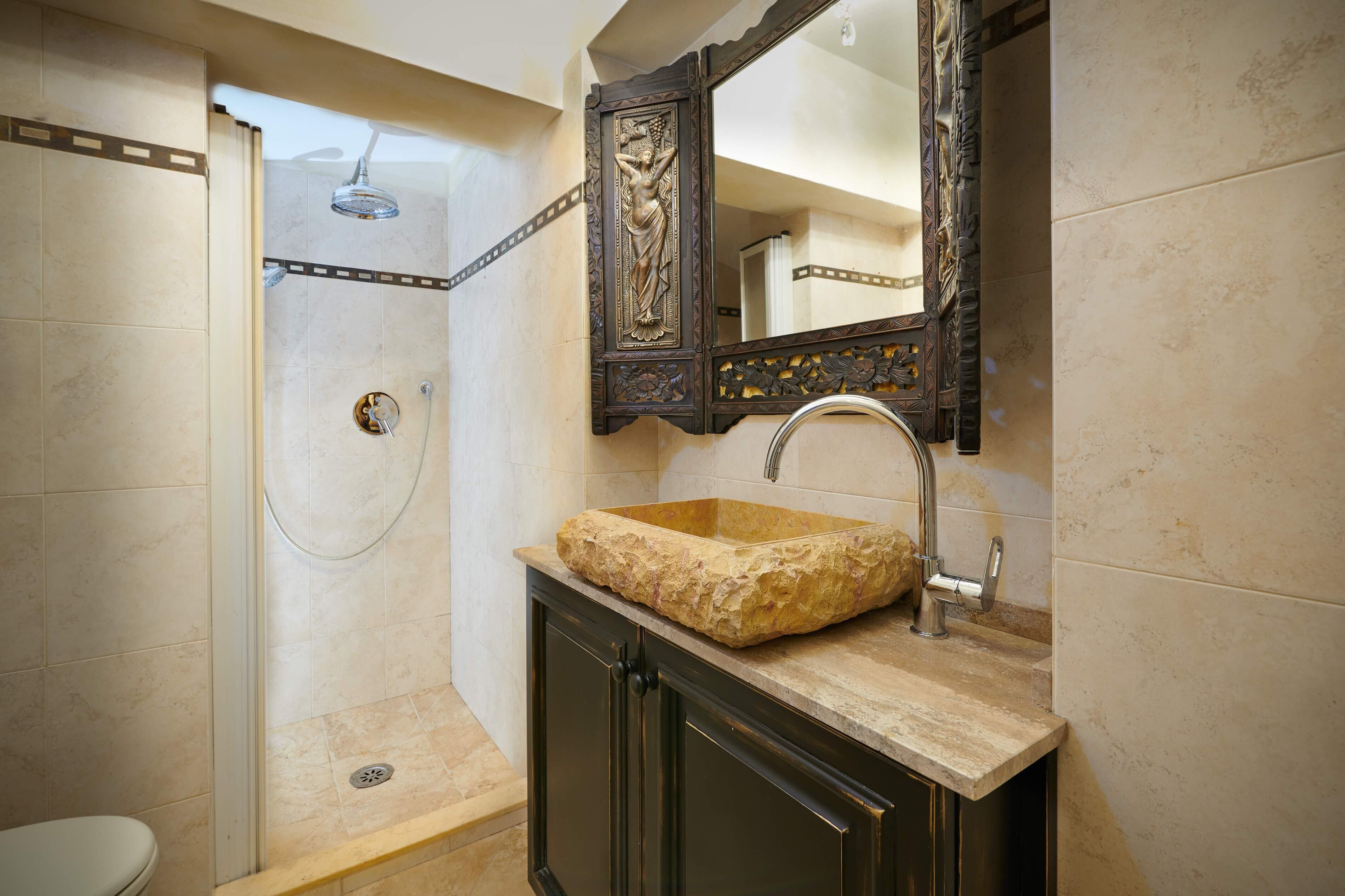 A bathroom with a stone sink, a shower, and a mirror with a decorative frame.