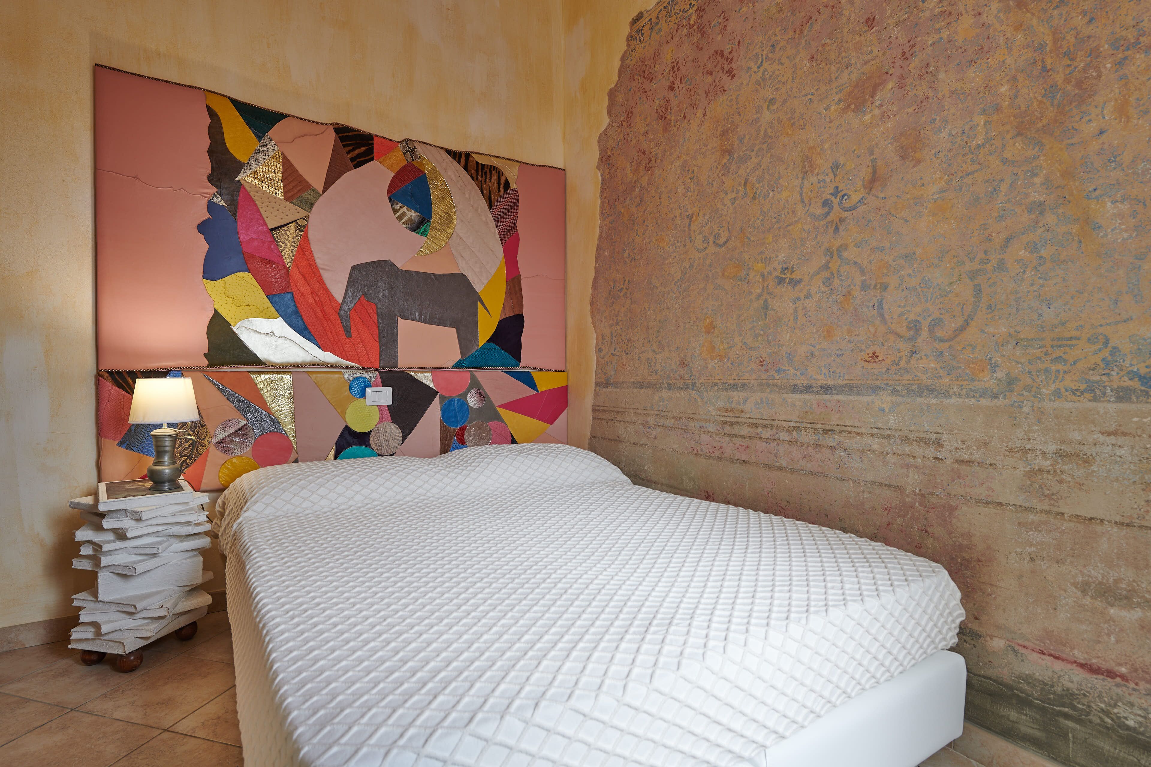 A bedroom with a bed, a stack of books, and a colorful abstract painting on the wall.