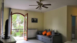Habitació amb un sofà gris, un ventilador de sostre, un radiador i una vista cap a un balcó per una porta oberta.
