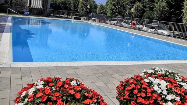 Una piscina amb una zona pavimentada amb rajoles, envoltada d'una tanca metàl·lica. Hi ha macissos de flors vermelles i blanques al primer pla.