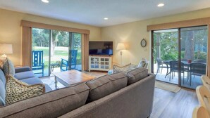 Un salón con sofá, sillones, una mesa de centro, televisor y un ventanal corredero que da a un espacio exterior con sillas.