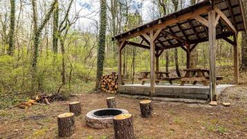 Un pabellón rústico al aire libre con una fogata y mesas de picnic rodeado de árboles.