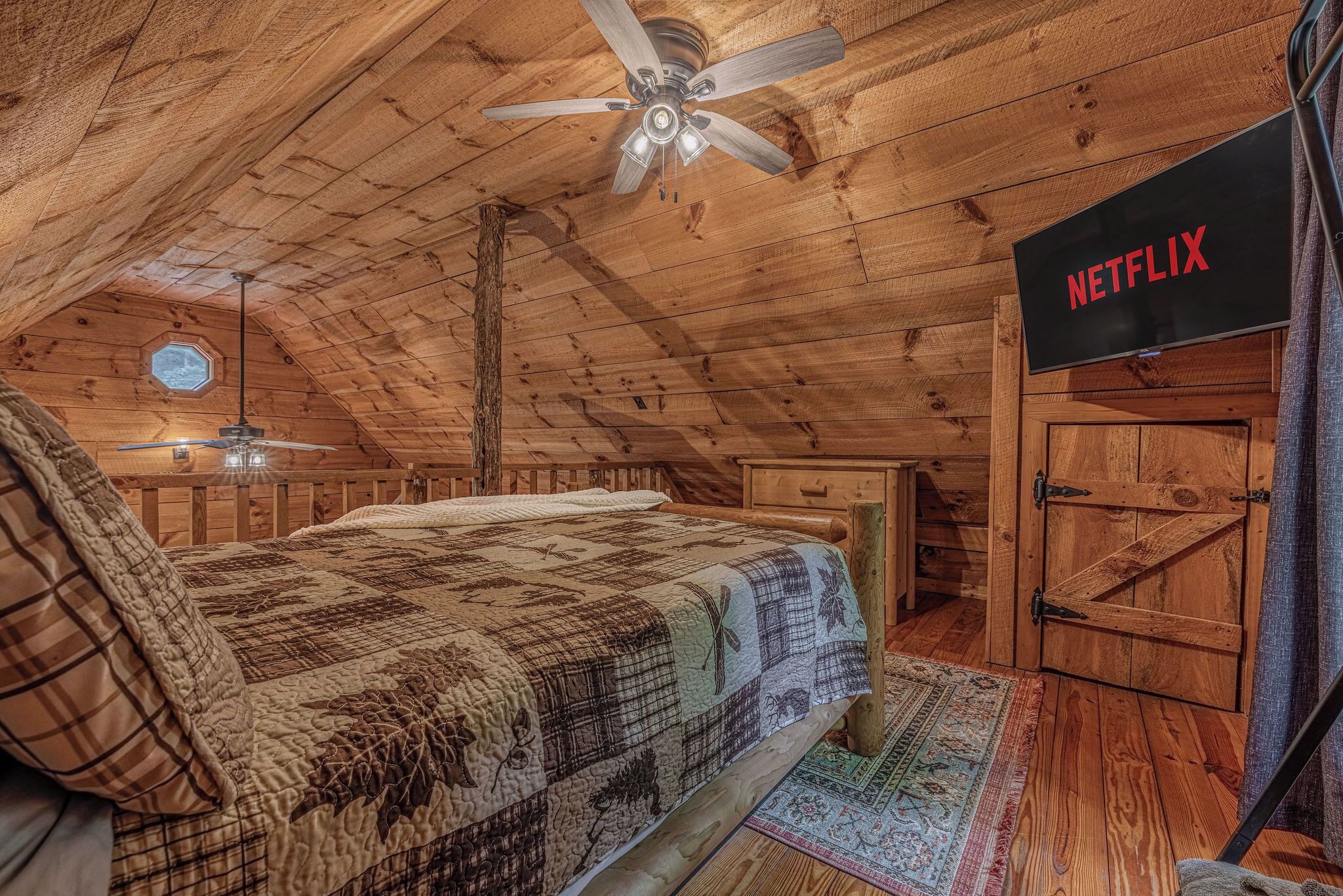 Ein gemütliches Schlafzimmer im Dachgeschoss mit Holzbalkendecke, einem Bett mit Steppdecke, einem Deckenventilator, einem Fernseher mit der Aufschrift 'NETFLIX' und einer rustikalen Holztür.