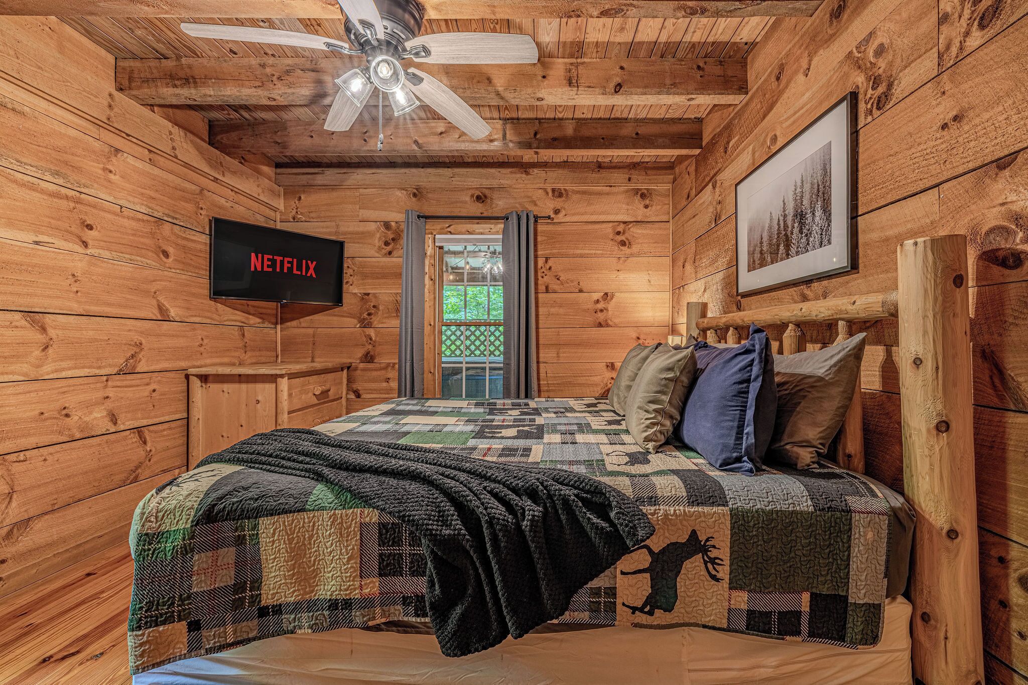 Camera da letto in una baita di legno, con un letto, una ventola a soffitto, un quadro incorniciato e un televisore che mostra 'NETFLIX'.