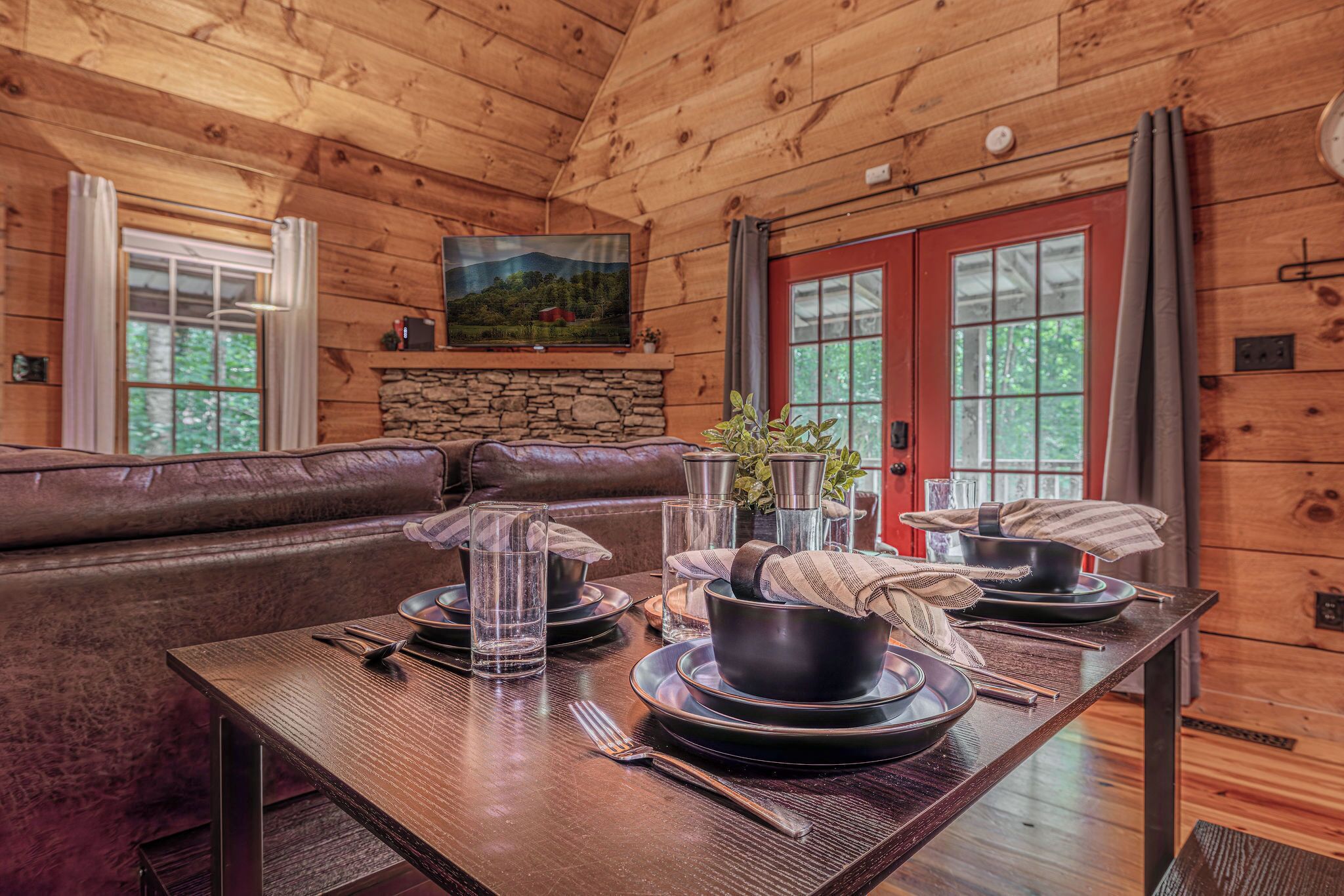 A wooden cabin interior with a dining table set for two, a stone fireplace, and a leather couch.