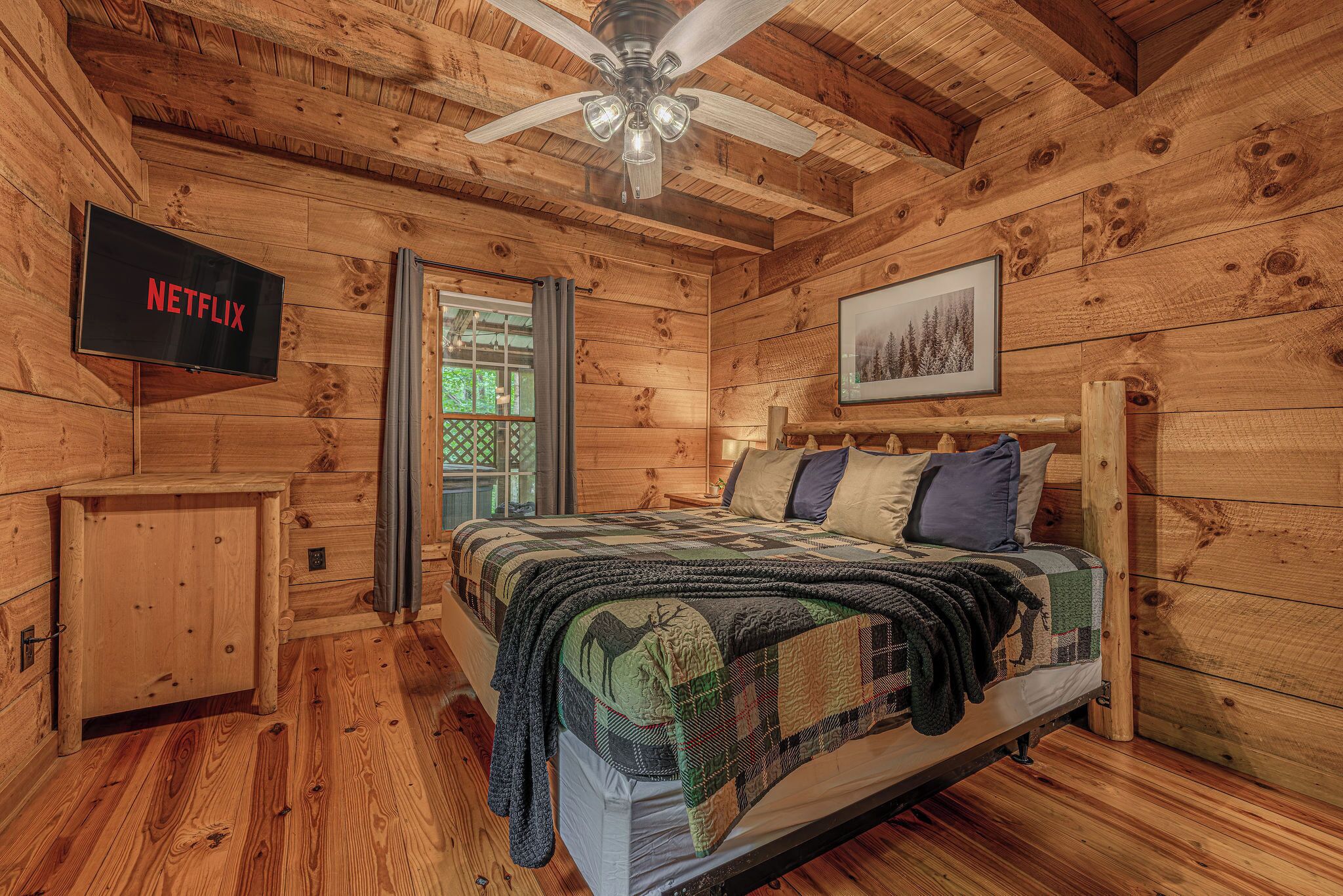 A wooden cabin room with a bed, a ceiling fan, a TV, and a door.