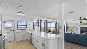 A modern kitchen with white cabinetry and a large island, a dining area with a blue sofa, and a living room with a fireplace. - Tipsy Turtle & Funky Pelican Massive Oceanfront (Palm Coast)
