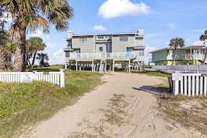 A beachfront vacation rental with a sandy path leading to a two-story building featuring balconies and a covered deck.