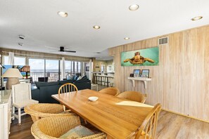 A spacious room with a wooden dining table, wicker chairs, a sofa, and a large window overlooking the ocean.