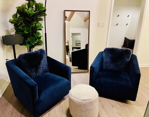 Quarto com duas poltronas azul-escuro com almofadas macias, um banquinho redondo, um espelho grande e um vaso de plantas.
