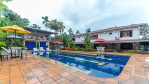 A swimming pool with lounge chairs and umbrellas, surrounded by palm trees and a building with a tiled roof. - Hotel Casa Baquero (Villavicencio)