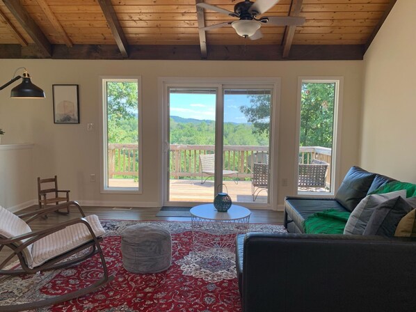 Ein gemütliches Wohnzimmer mit Holzbalkendecke, Deckenventilator, einer großen Schiebeglasdoor zum Außenbereich und einer Terrasse, einer schwarzen Ledersofa, einem Schaukelstuhl und einem gemusterten Teppich.