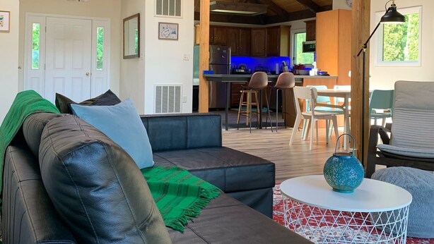Un salón espacioso con techo alto y vigas de madera a la vista, un sofá de cuero oscuro, una mesa de centro redonda y una zona de cocina al fondo.