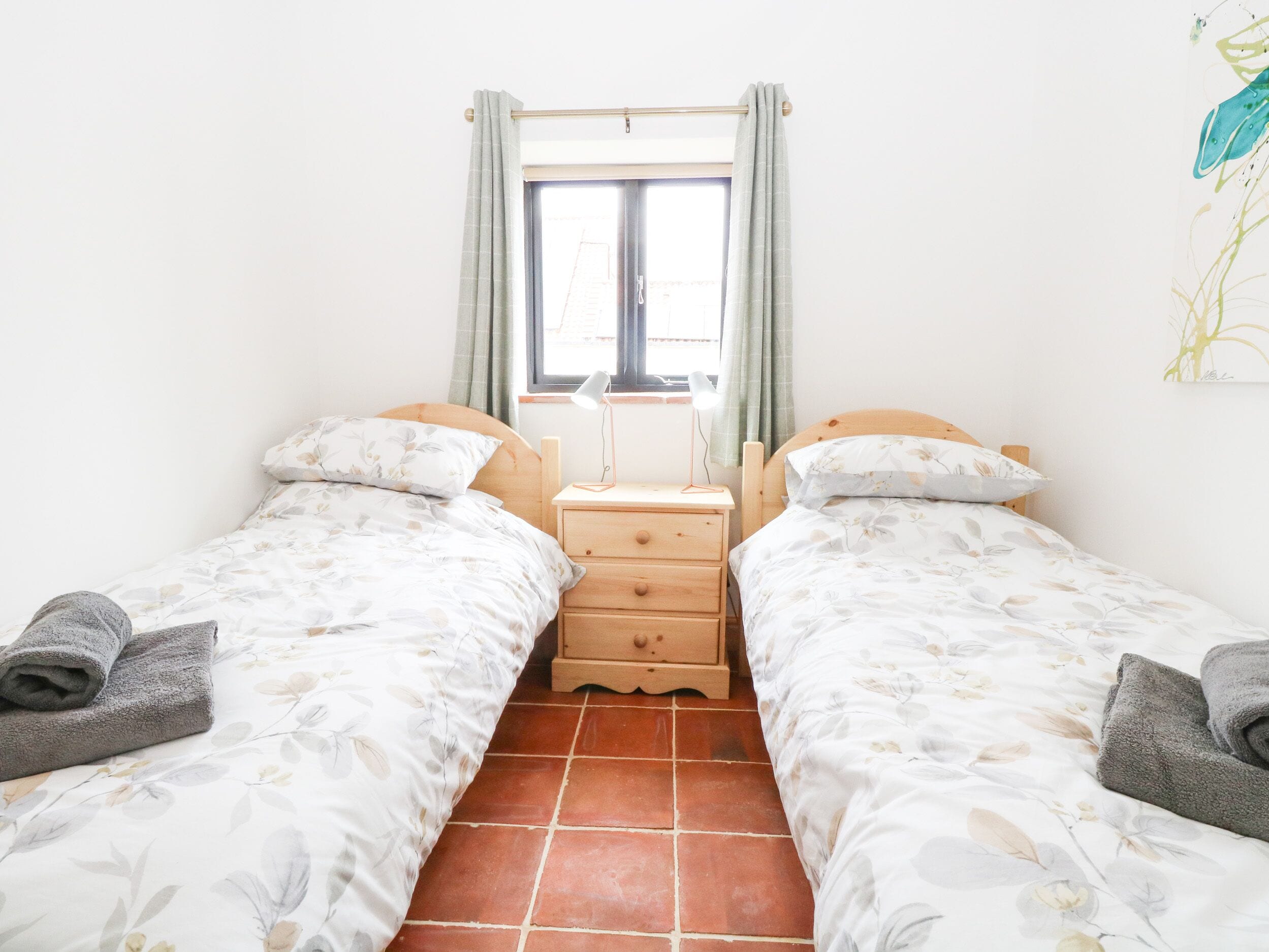 Habitación doble con dos camas, una ventana con cortinas, una mesita de noche con lámpara y suelo de baldosas.