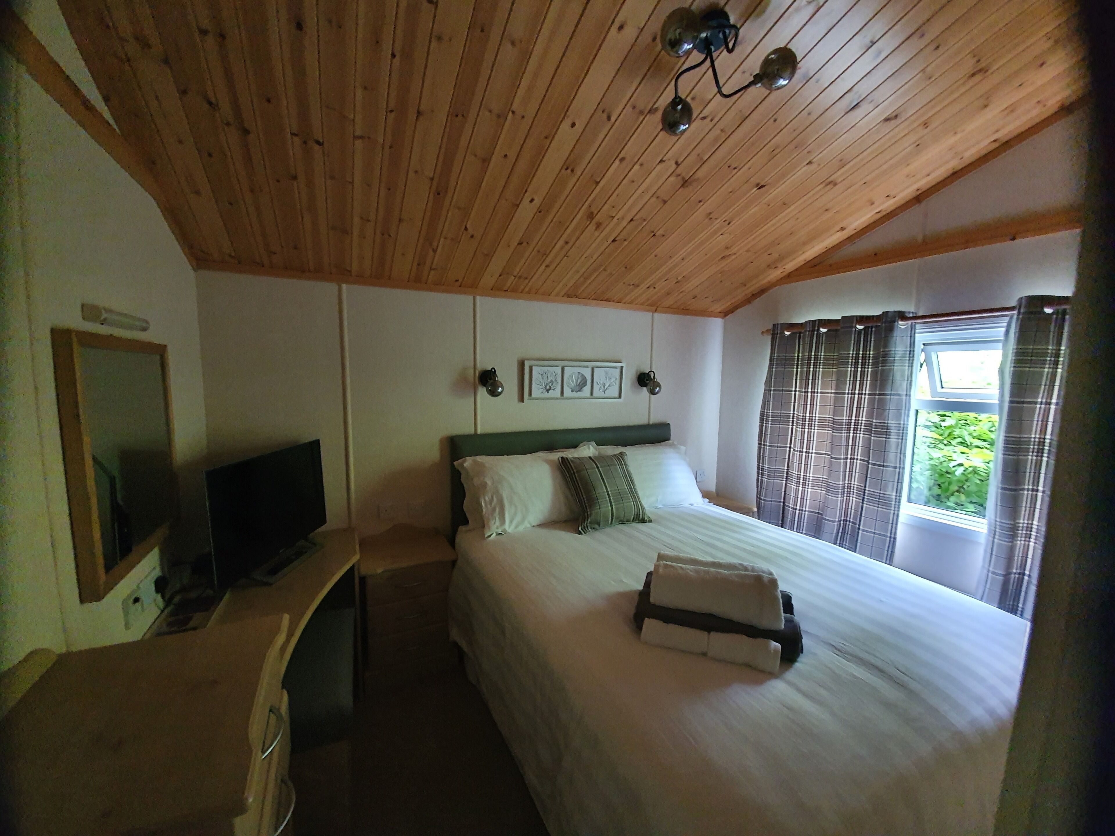 Une chambre avec un plafond en bois, un lit avec une literie blanche, une table de chevet avec une télévision et une fenêtre avec des rideaux.